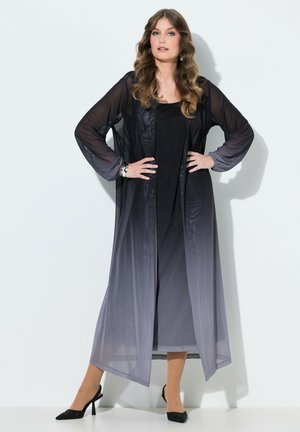 Femme aux cheveux ondulés portant un long cardigan léger ombré noir-gris sur une robe noire et des talons noirs, debout les mains sur les hanches.