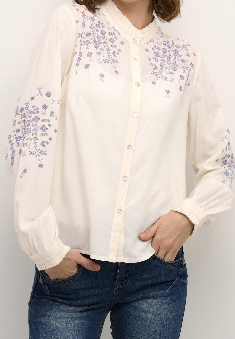 Blouse crème avec broderies florales en bleu et orange, devant boutonné, manches longues avec poignets froncés, tissu léger.