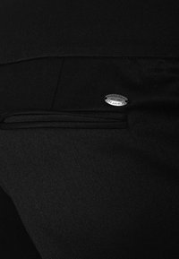 Zwarte broek met een zijzak en een zilveren merklabel met de tekst "GUESS EST. 1981 LOS ANGELES," tentoongesteld op een textiel met een structuur.