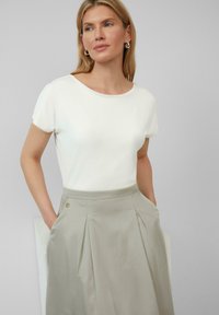 Wit kort mouwen top gecombineerd met een lichtgrijze geplooide rok met zijzakken en een gouden accentknop. Gladde texturen overal.