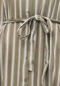 Φόρεμα πουκάμισο με κάθετες λευκές ρίγες σε ελιά πράσινο ύφασμα, με ζώνη από το ίδιο ύφασμα δεμένη στη μέση.