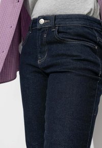 Jean en denim foncé avec une coupe slim, poches avant et fermeture à bouton, avec des coutures contrastées le long des coutures.