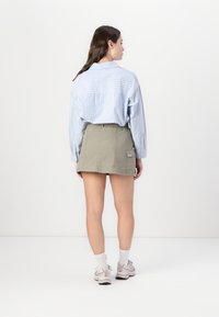 Camicia over-size a righe blu chiaro e bianco con bottoni; gonna verde oliva, lunga fino alla coscia, con vita elasticizzata e tasca laterale.