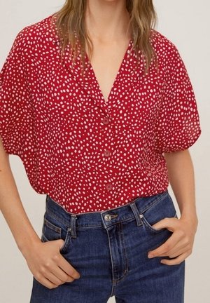 Kvinde iført en rød kortærmet bluse med hvide prikker og knapper, stoppet ned i højtaljede blå jeans med hænderne i lommerne.