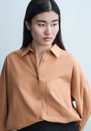 Jeune femme aux longs cheveux noirs portant une chemise ample couleur beige à boutons, regardant sur le côté contre un fond clair uni.