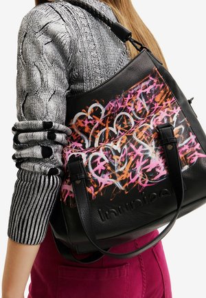 Sac fourre-tout en cuir noir avec des graffitis de cœurs colorés en rose, orange et blanc, porté à l'épaule par-dessus un pull en maille gris et un pantalon violet.