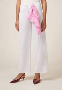 Femme portant un pantalon blanc à jambes larges avec une écharpe à motifs roses nouée à la taille et des escarpins bordeaux à bout pointu.
