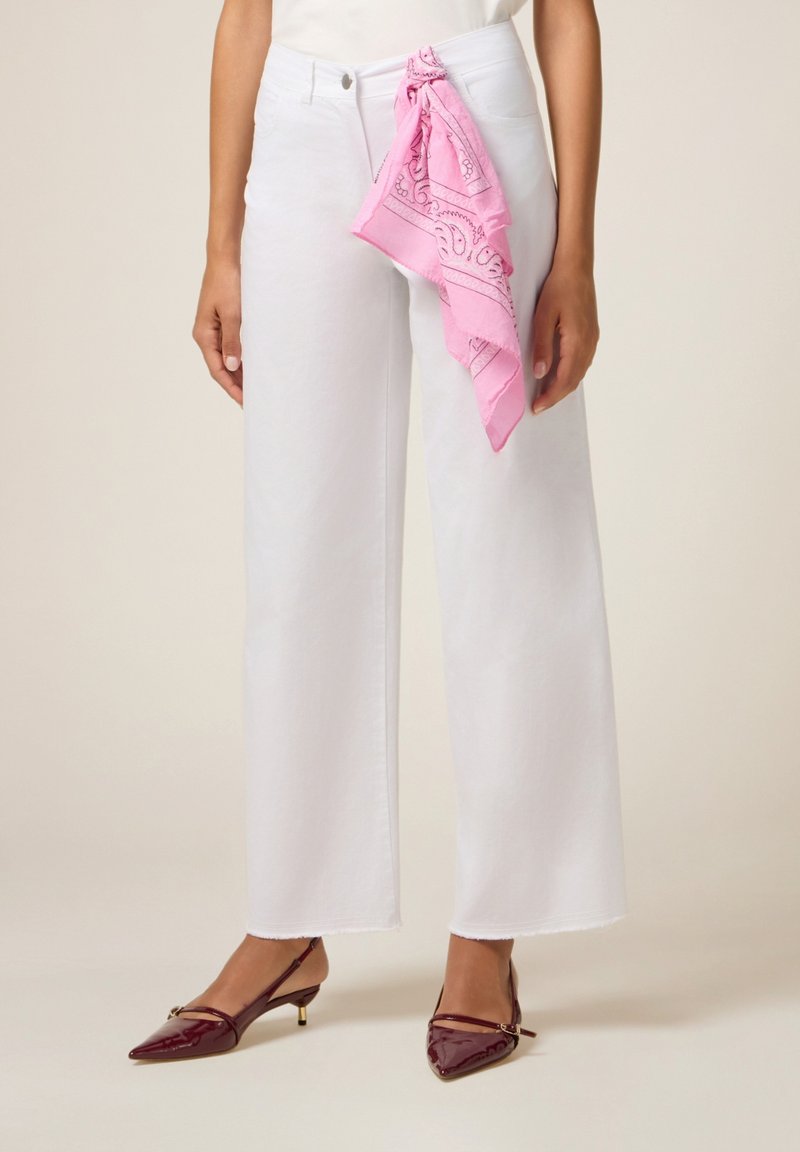 Femme portant un pantalon blanc à jambes larges avec une écharpe à motifs roses nouée à la taille et des escarpins bordeaux à bout pointu.