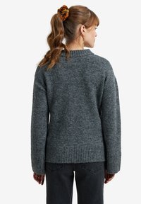 Frau mit braunen Haaren im Pferdeschwanz, die einen grauen Strickpullover und schwarze Jeans trägt und mit dem Rücken zur Kamera steht.