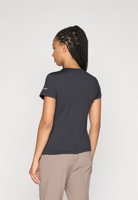Kortsleeve svart T-skjorte laget av glatt stoff, med rund hals og minimal logo på ermet, kombinert med beige bukser.