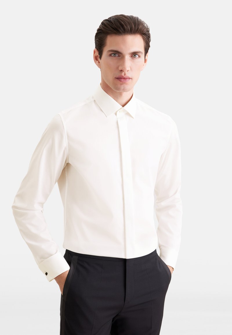 Homme aux cheveux foncés portant une chemise blanche et un pantalon noir, debout les mains dans les poches devant un fond clair uni.