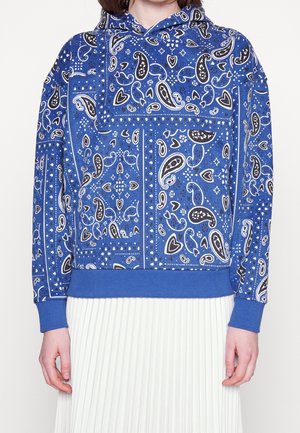 Blaue Kapuzenjacke mit einem Schwarz-Weiß-Paisleymuster, versehen mit einer Kordelzugkapuze und gerippten Bündchen, kombiniert mit einem plissierten weißen Rock.