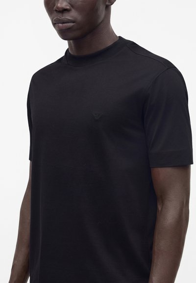 Μαύρο κοντομάνικο T-shirt με στρογγυλή λαιμόκοψη που φοράει ένα άτομο, με μικρό, διακριτικό έμβλημα στο αριστερό στήθος, σε απλό λευκό φόντο.