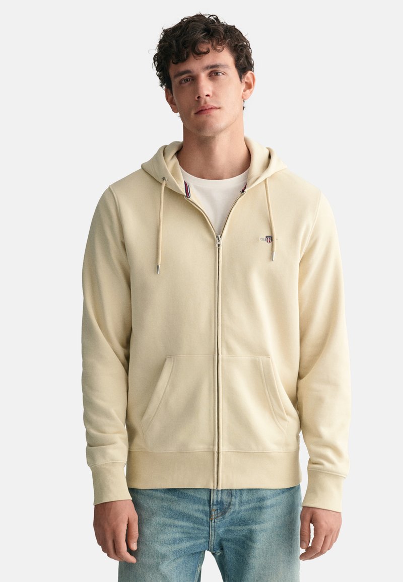 Beige Kapuzenjacke mit Reißverschluss, Fronttaschen, verstellbarem Kapuzenzug und gerippten Bündchen. Weicher Stoff mit dezentem Logo und buntem Krageninnenfutter.