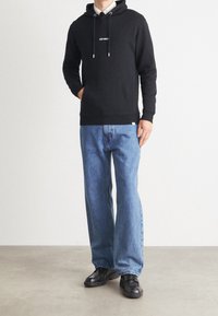 Czarna bluzka z kapturem z białym napisem "LES DEUX," w połączeniu z jasnoniebieskimi prostymi dżinsami i czarnymi butami. Minimalistyczny design i luźny krój.