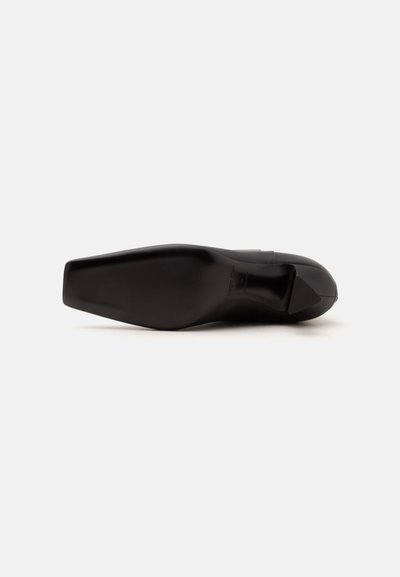 Chaussure habillée en cuir noir vue par la semelle, montrant une forme d'orteil carrée et un talon bas sur fond blanc.