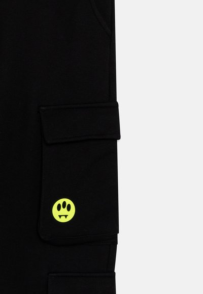Pantaloni jogger neri realizzati in tessuto morbido, con una tasca laterale e una grafica di una faccina sorridente gialla con i canini. Design semplice con una cintura a coste aderente.