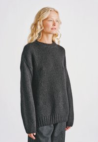 Blondynka z falującymi włosami, ubrana w luźny, ciemnoszary sweter z dzianiny oraz pasujące szare spodnie, stojąca na tle jasnego, jednolitego tła.