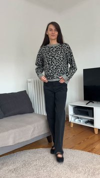 Sivi džemper s crnim leopardskim uzorkom, rebrasti manšeti, upareni s crnim hlačama širokih nogavica. U pozadini su kauč i televizor.