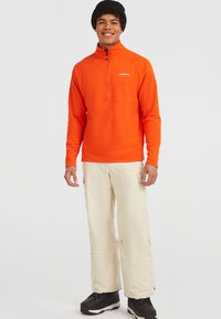 Felroze oranje pullover met een halve ritssluiting, voorzien van lange mouwen en een ontspannen pasvorm, gecombineerd met crèmekleurige cargobroeken en zwarte laarzen.