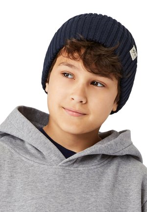 Marineblå ribbet strikket hue med et mærke, båret af et barn. Grå hoodie under, der viser hætten og dele af en marineblå t-shirt. Nærbillede af ansigtet.