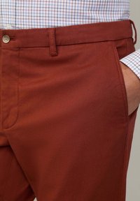 Close-up van een persoon die roestkleurige broek draagt met een geknoopte tailleband en een hand in de rechterzak, gecombineerd met een geruit shirt.