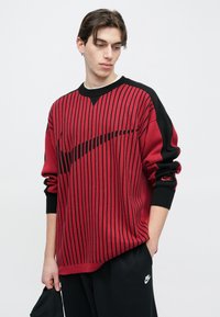 Piros és fekete csíkos kötött pulóver bordázott, kerek nyakkivágással és fekete mandzsettákkal. Közepén nagy fekete Nike logó található.