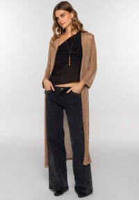 Cárdigan largo de punto en tono bronce metálico, con el frente abierto; combinado con una blusa negra y unos jeans negros de pierna ancha con adornos de tachuelas en el dobladillo.