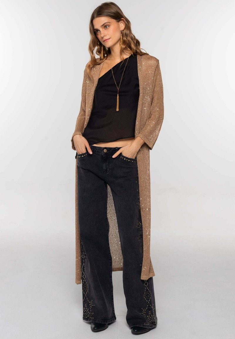 Cárdigan largo de punto en tono bronce metálico, con el frente abierto; combinado con una blusa negra y unos jeans negros de pierna ancha con adornos de tachuelas en el dobladillo.