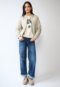 Veste bomber matelassée beige clair, pull graphique crème, jean denim bleu décontracté roulé aux chevilles, chaussures beige à enfiler. Palette de couleurs neutres.