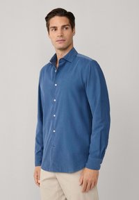 Camicia blu a maniche lunghe in tessuto testurizzato, con colletto a bottoni e bottoni bianchi, orlo curvo e design aderente.