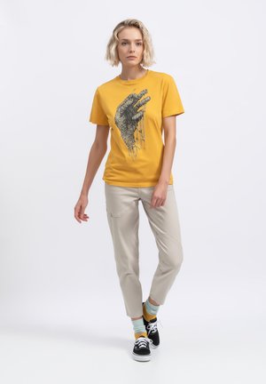T-shirt en coton jaune avec un graphique représentant une main détaillée ; associé à un pantalon beige et des baskets noires. Le mannequin se tient devant un fond blanc.