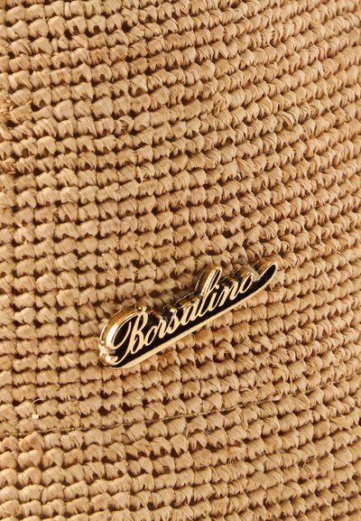 Κοντινή λήψη υφής πλεγμένου άχυρου με χρυσή και μαύρη μεταλλική ετικέτα «Borsalino» προσαρτημένη.