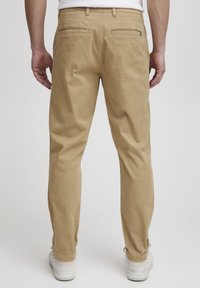 Chino kaki en coton, à coupe droite, avec deux poches arrière et une fermeture éclair. Une paire de baskets blanches visible en bas.