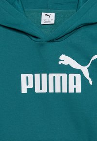 Φούτερ με κουκούλα σε τιρκουάζ χρώμα με μεγάλο λευκό λογότυπο Puma και κείμενο στο μπροστινό μέρος, που δείχνει την ετικέτα μεγέθους μέσα στον γιακά: ΗΠΑ/ΚΑ M, ΗΒ 11-12 ετών, Γερμανία/Ιταλία 152.