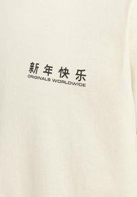 Balta medvilnė marškinėlių su juodu grafiniu teksto dizainu, kuriame yra kinų ženklai ir frazė "ORIGINALS WORLDWIDE." Glotni tekstūra.