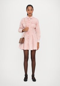Roze denim jurk met lange mouwen, knoopsluiting aan de voorkant, twee borstzakken, uitlopende rok, gecombineerd met zwarte platte schoenen en een bruine crossbody tas.