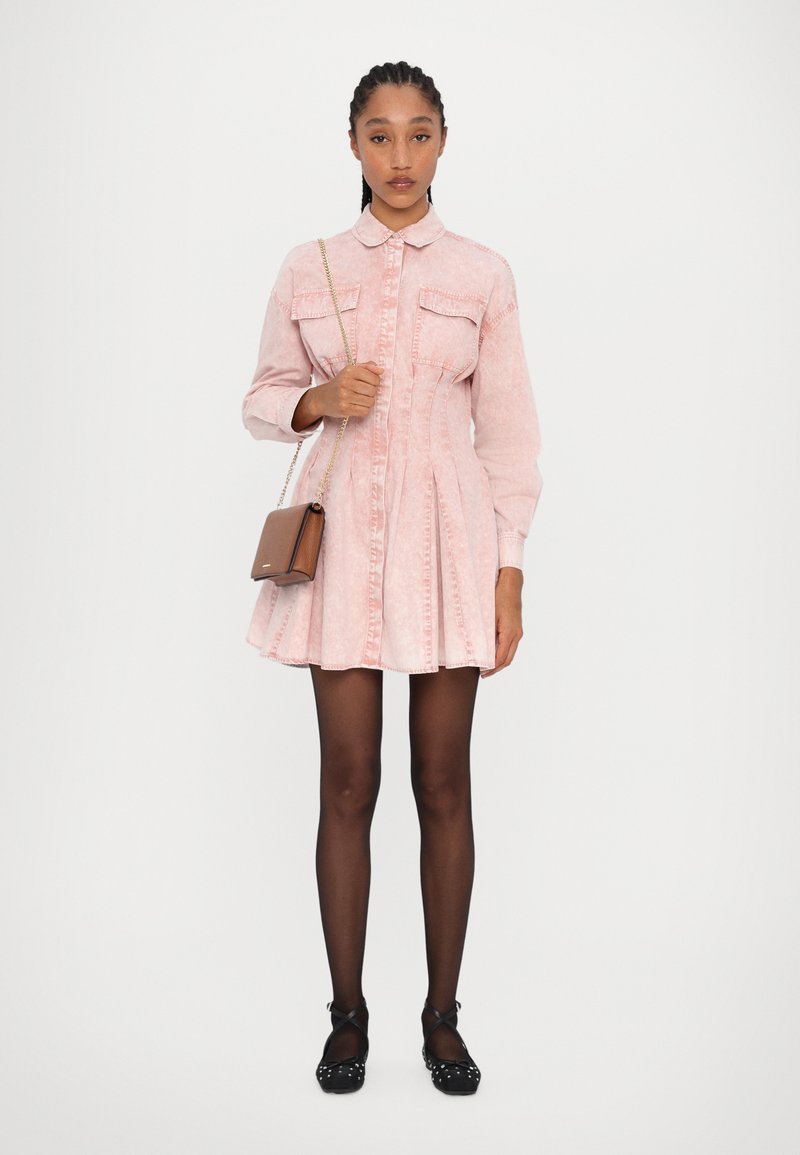 Roze denim jurk met lange mouwen, knoopsluiting aan de voorkant, twee borstzakken, uitlopende rok, gecombineerd met zwarte platte schoenen en een bruine crossbody tas.