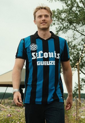 Blauw en zwart gestreepte voetbalshirt met kraag, met witte letters "St. Louis" en "Gueuze." Model draagt een donkere spijkerbroek.