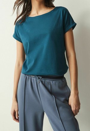 Teal cotton t-shirt with rolled sleeves, paired with loose-fitting gray pants featuring a drawstring waistband and metal accent detail.