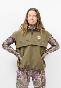 Olivgrüner Fleece-Pullover mit Frontreißverschluss und aufgesticktem Text. Die innere Schicht besteht aus einem gemusterten, mehrfarbigen Langarmoberteil und Leggings.