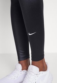 Černé legíny s lesklou texturou a bílým logem Nike blízko kotníku, spárované s bílými sportovními botami s prodyšným designem.