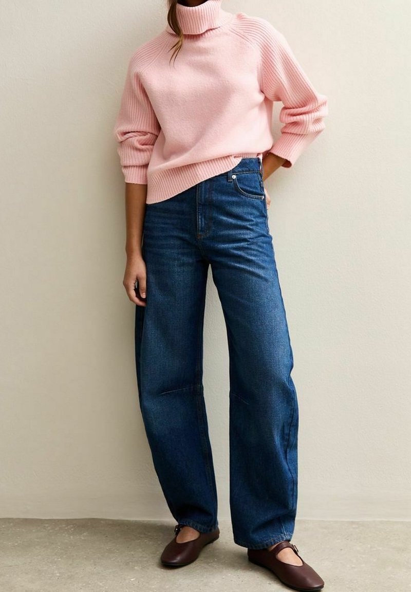 Femme portant un pull à col roulé rose clair, un jean bleu à taille haute et jambe large, et des chaussures plates marron foncé, debout contre un mur uni.