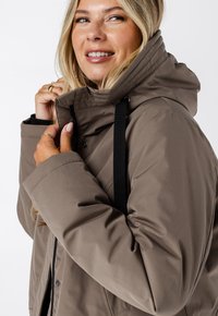 Donkerbruine geïsoleerde jas met een hoge kraag, voorzien van een rits- en drukknoopsluiting, zwarte koordjes en een gladde, matte textuur.