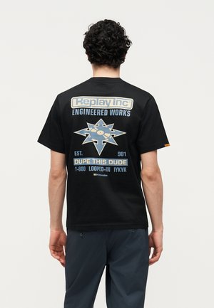 Mann mit lockigem Haar, trägt ein schwarzes Replay Inc T-Shirt mit blauem und beigem Stern-Logo und Text auf dem Rücken, dunkle Hose, von hinten gesehen.