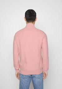 Ljusrosa stickad tröja med hög krage, ribbade ärmslut och nederkant. Tyget verkar mjukt och texturerat, med en enkel design.