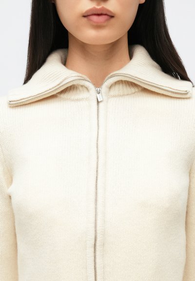 Kvinne iført en kremfarget, strikket genser med glidelås og bred krage, med langt, rett, mørkt hår som er synlig ved skuldrene.