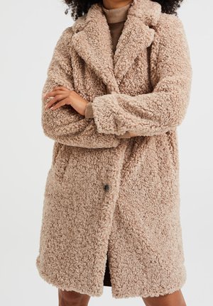 Femme portant un manteau teddy beige mi-long avec un large col et un tissu texturé bouclé sur un pull à col roulé marron clair.