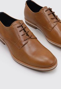 Chaussures habillées en cuir marron avec une finition lisse, bout arrondi, six œillets et une semelle blanche contrastante. Design minimaliste et silhouette élégante.