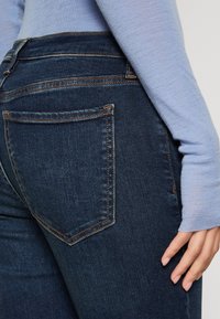 Mörkblå denimjeans har hög midja, femfickorsdesign, synlig sömnad och en slät textur. En hand vilar på höfterna.